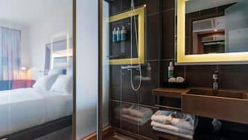 A modern hotel room with a glass partition, a large bed, a shower area, and a bathroom with a mirror and towel rack.