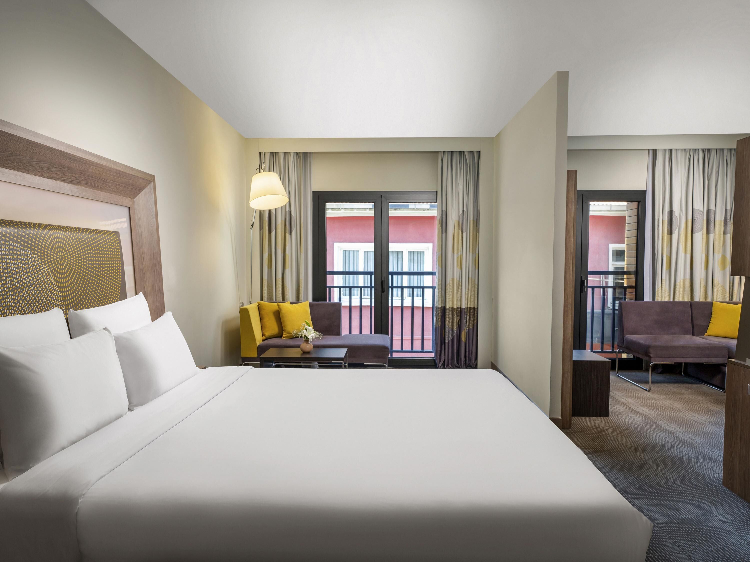 A hotel room with a large bed, a sofa, a small table, and a chair. There is a balcony with curtains and a view of the outside.