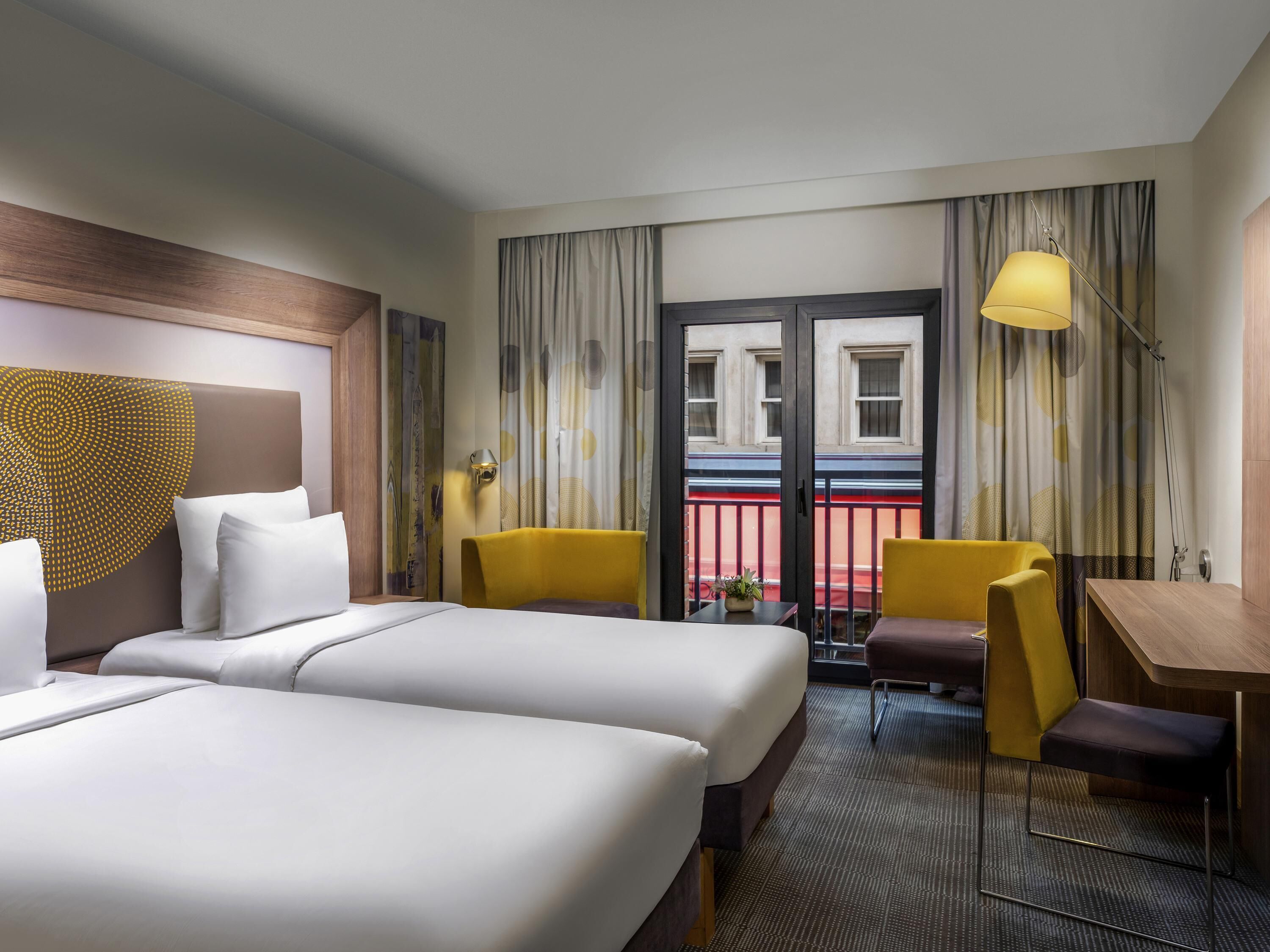 A hotel room with two beds, a desk, a chair, a lamp, and a view of the street through a door.