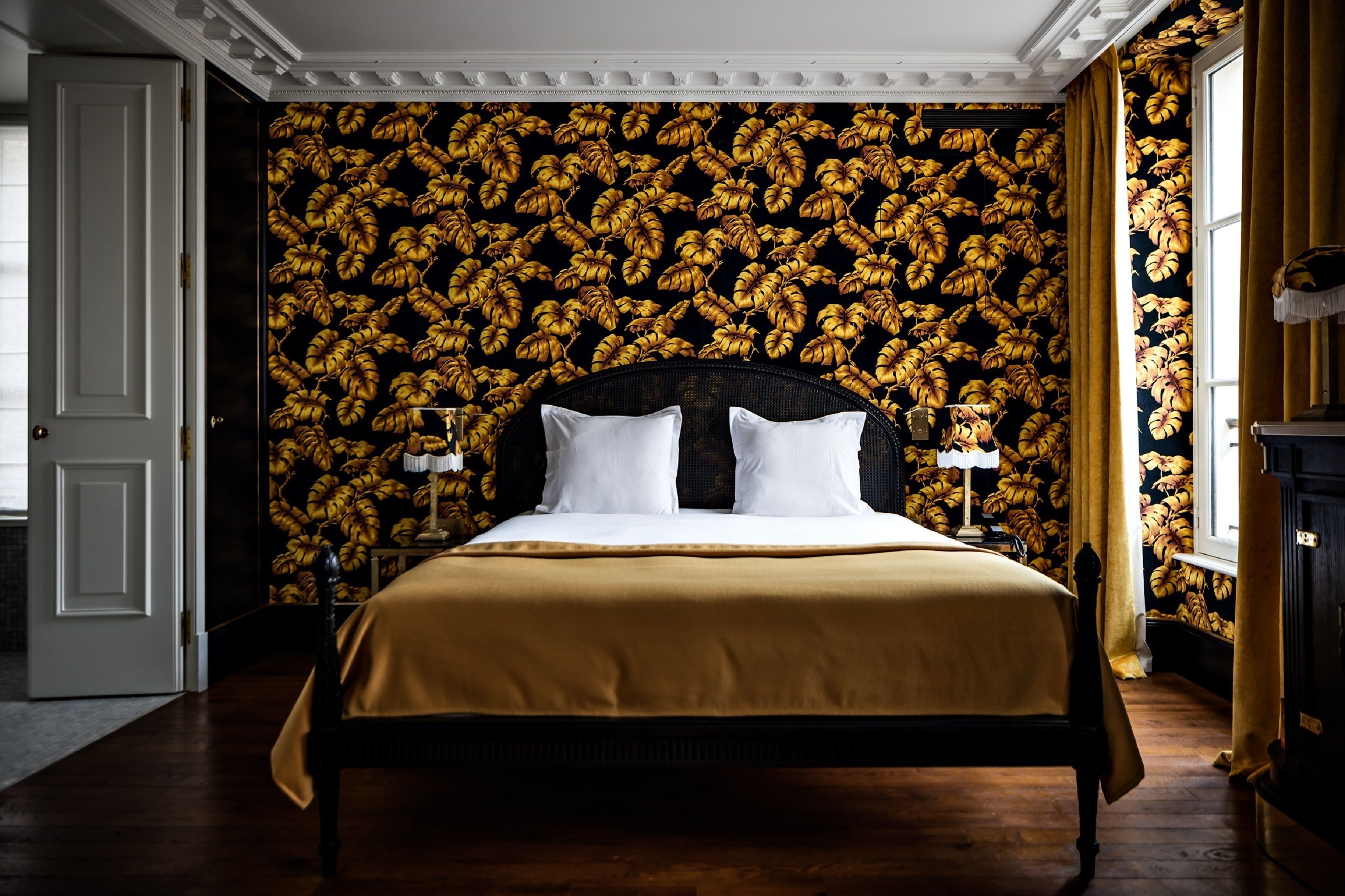 Una cama con cabecera oscura, almohadas blancas y cubrecama dorado en una habitación con papel tapiz estampado y suelo de madera.