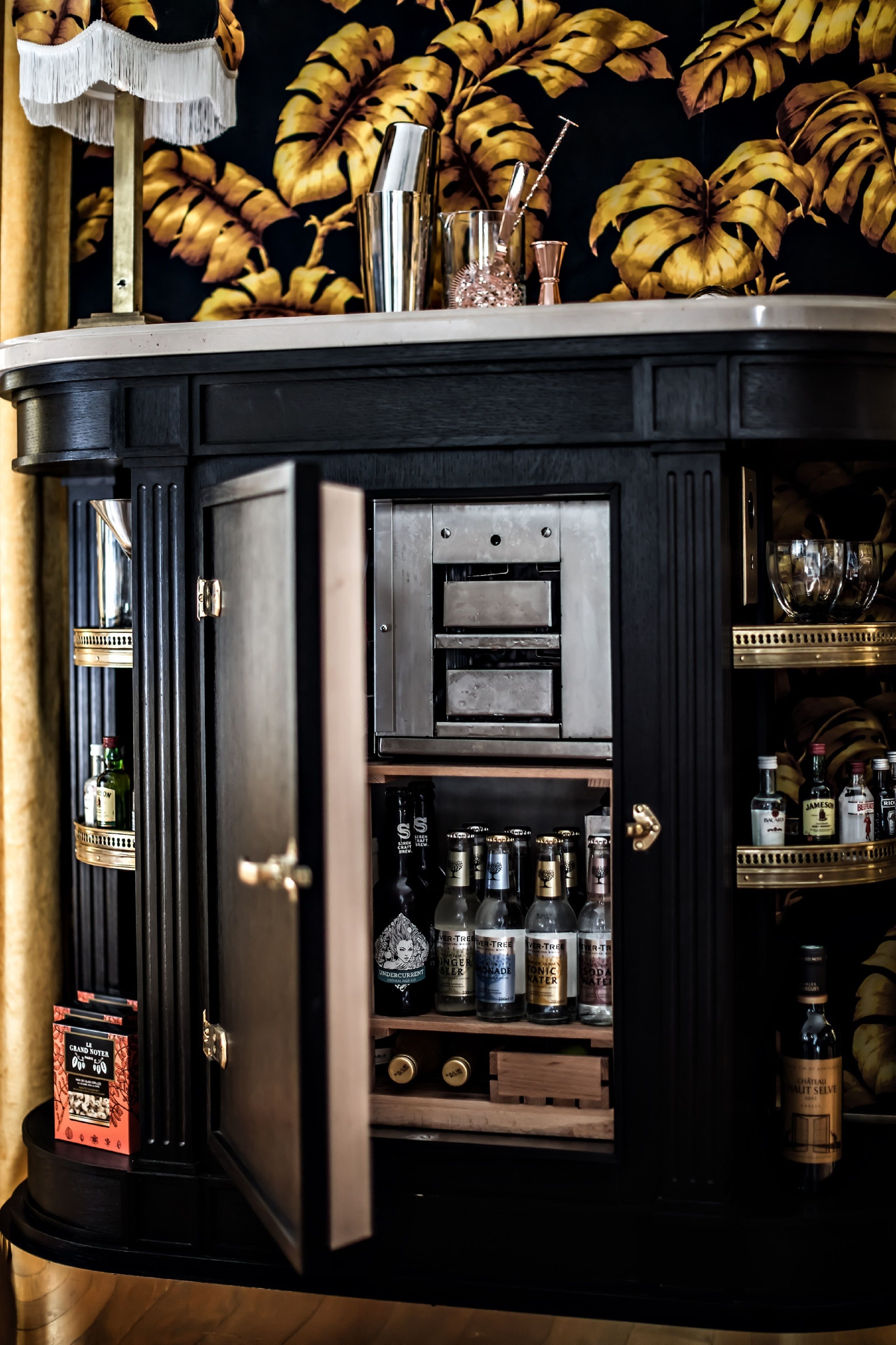Uma área de bar com armário de madeira escura, prateleiras com garrafas e um arranjo decorativo de plantas.