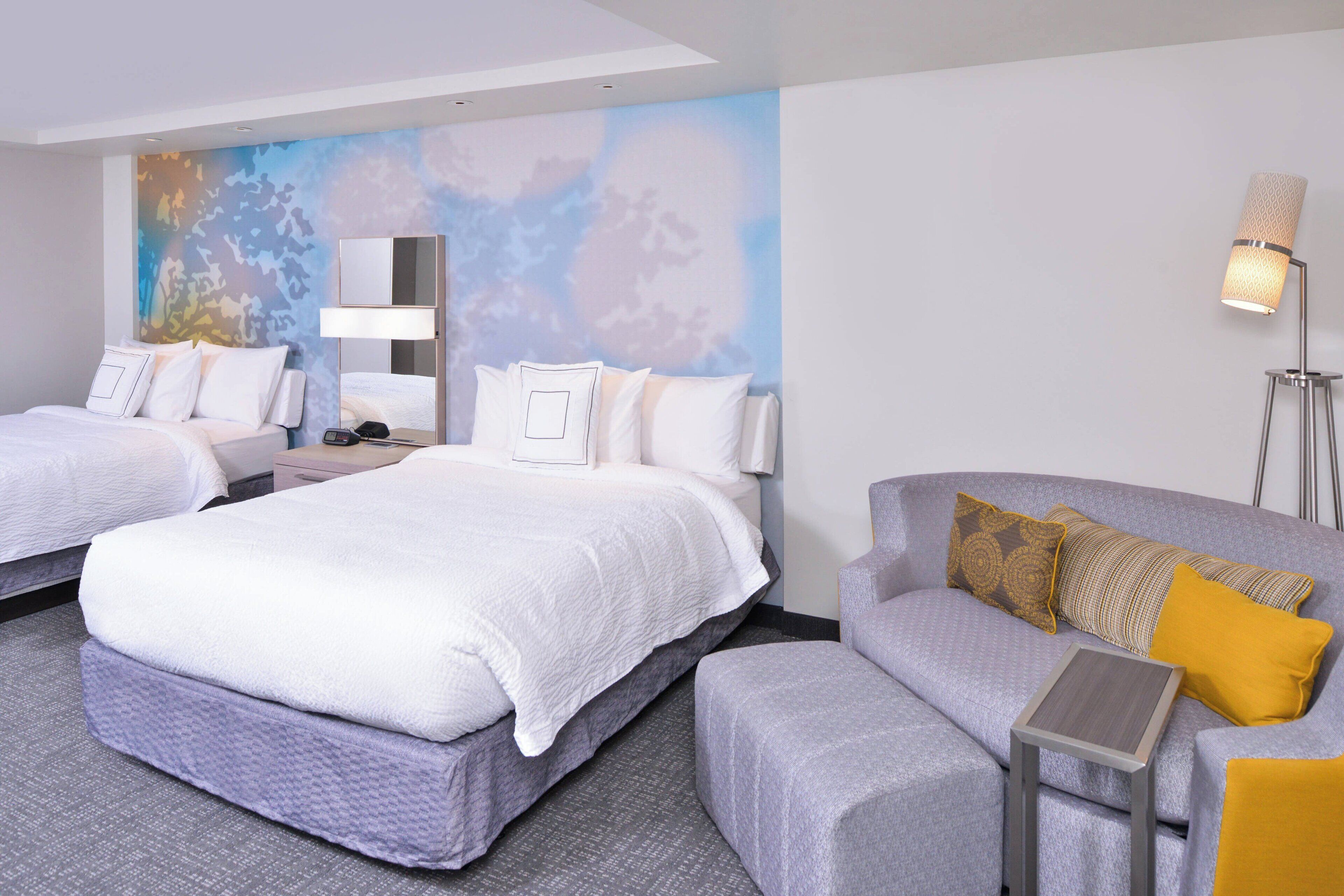 Quarto de hotel com duas camas, um sofá, uma mesinha e um abajur.