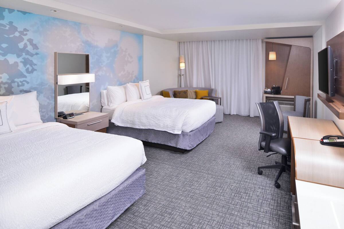 A hotel room with two beds, a desk, a TV, and a desk lamp.