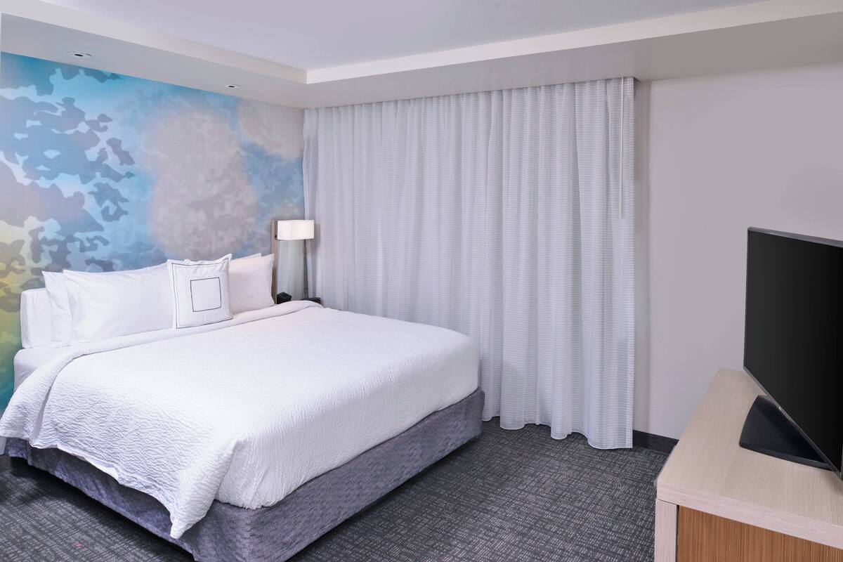 A hotel room with a bed, a flat-screen TV, a wall-mounted lamp, and a large window with curtains.