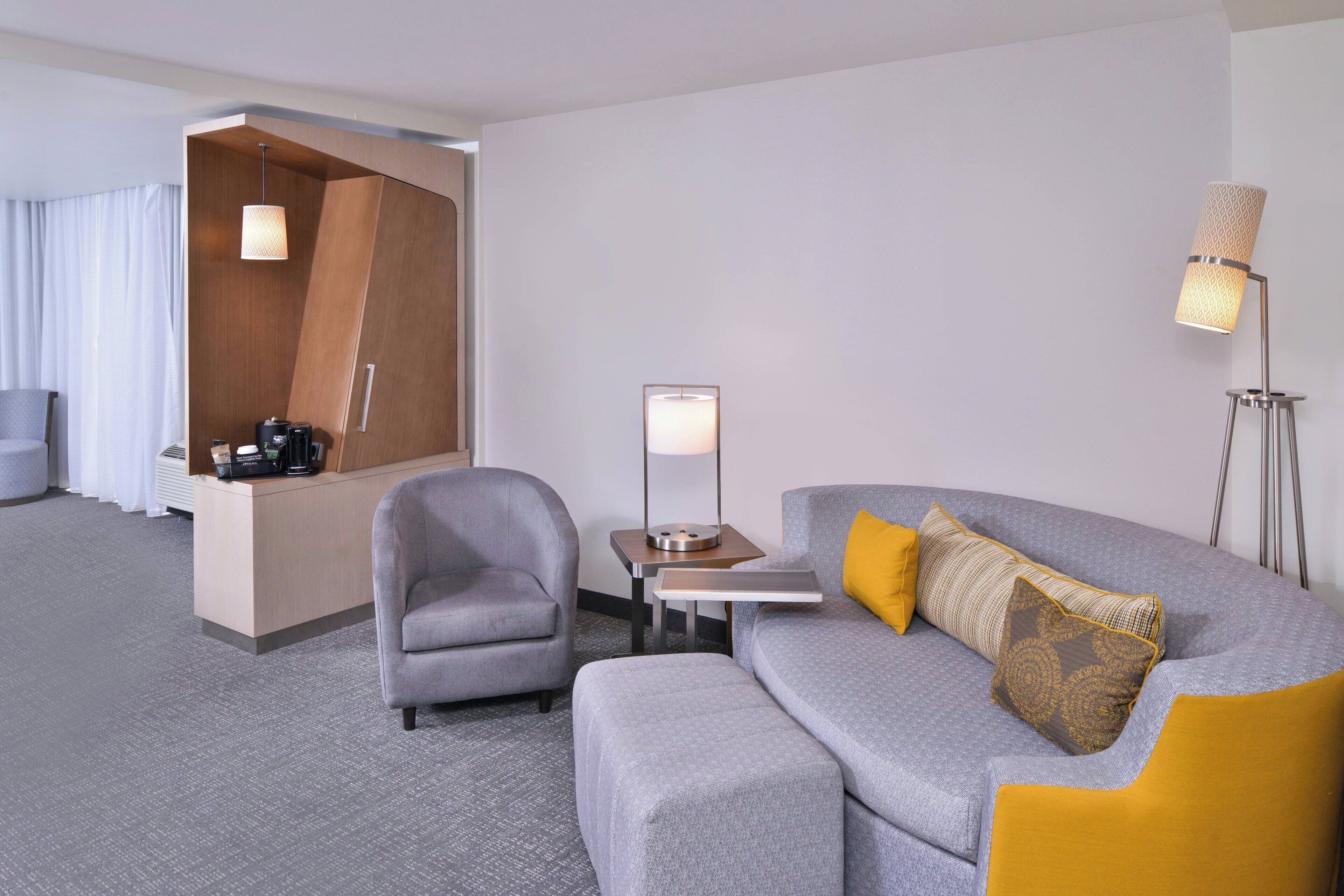 Una habitación de hotel moderna con un sofá gris, un sillón gris, una mesita con una lámpara y un escritorio con una cafetera.