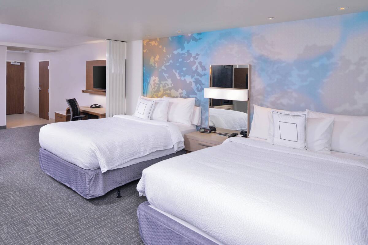 A hotel room with two beds, a desk, a chair, a TV, and a wall with a blue and white abstract design.
