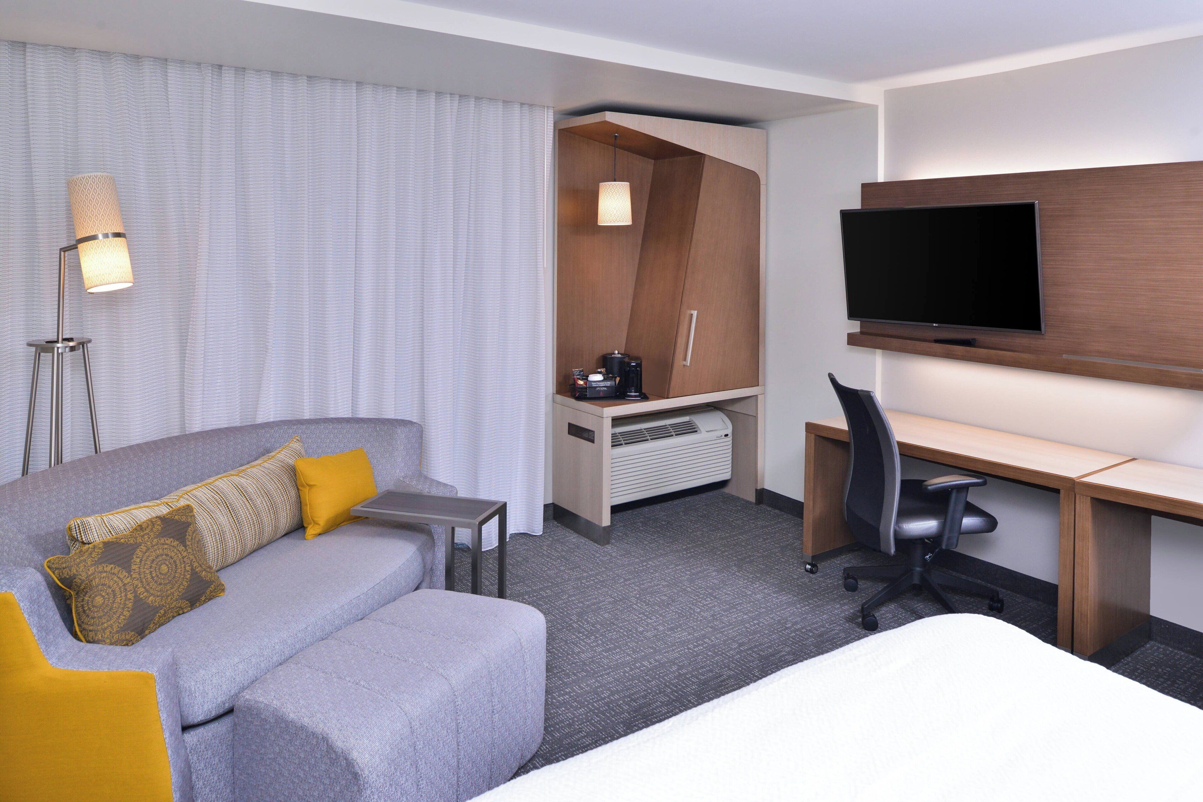 Una habitación de hotel con un sofá gris, un escritorio pequeño, una silla, un televisor de pantalla plana y una cama.