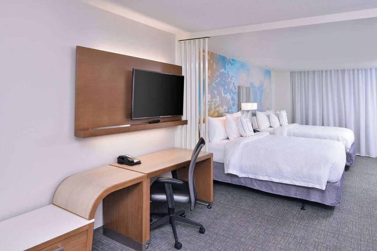 A hotel room with a desk, two beds, a TV, and a large window with curtains.