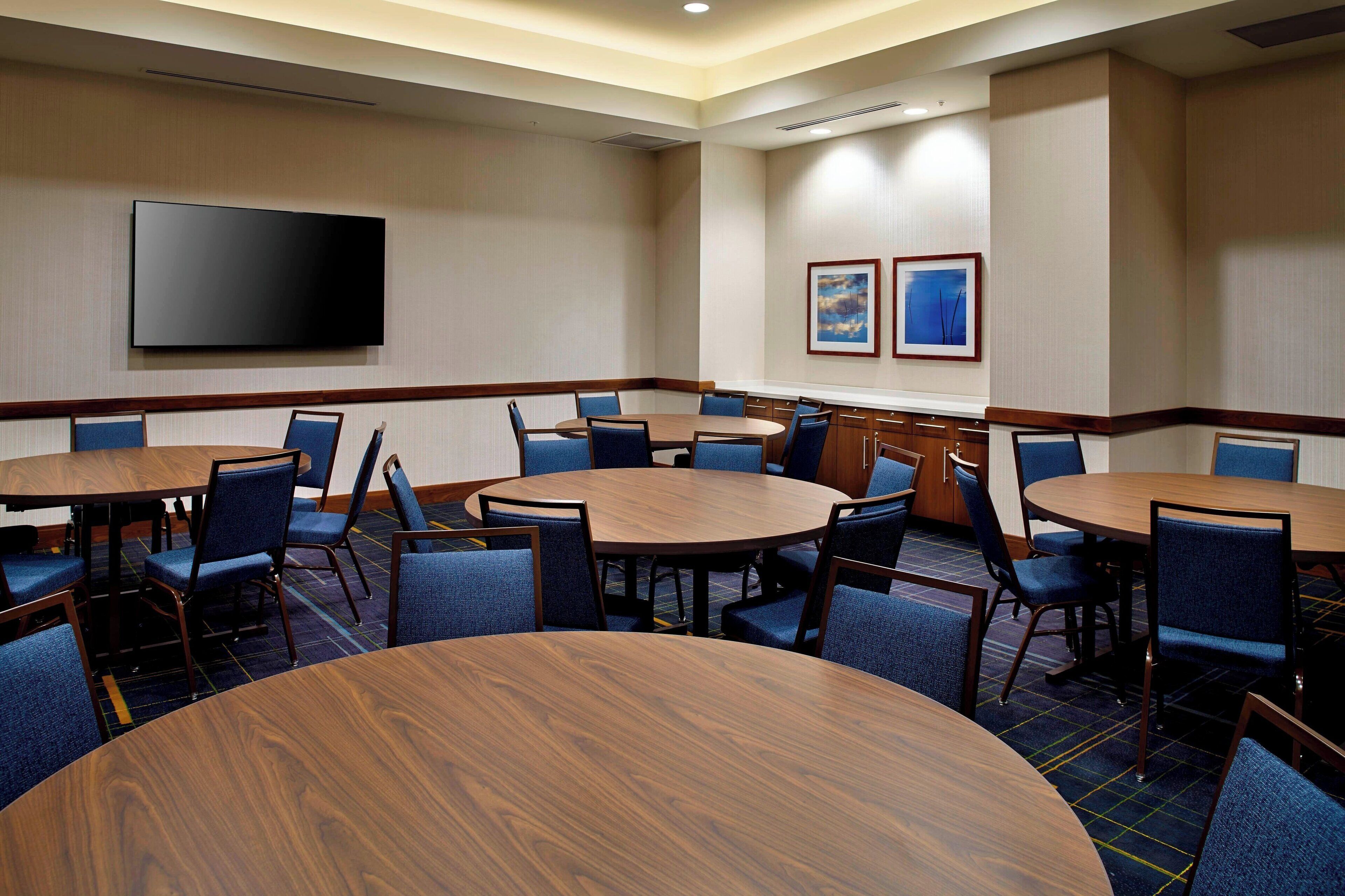 A conference room with multiple round tables and chairs, a flat-screen TV mounted on the wall, and two framed pictures on the wall.