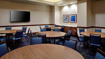 A conference room with multiple round tables and chairs, a flat-screen TV mounted on the wall, and two framed pictures on the wall.