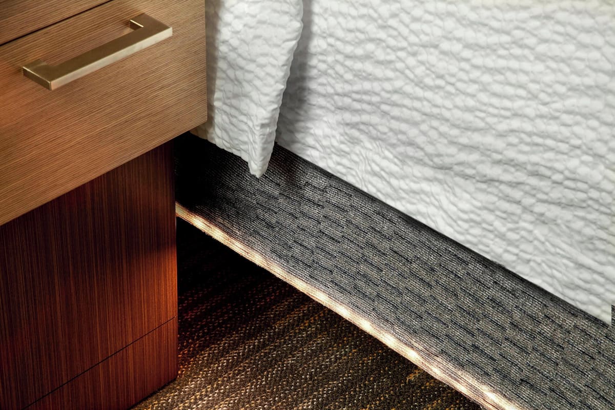 Un tiroir en bois avec une poignée métallique, un couvre-lit blanc texturé et un tapis à motifs sombres.