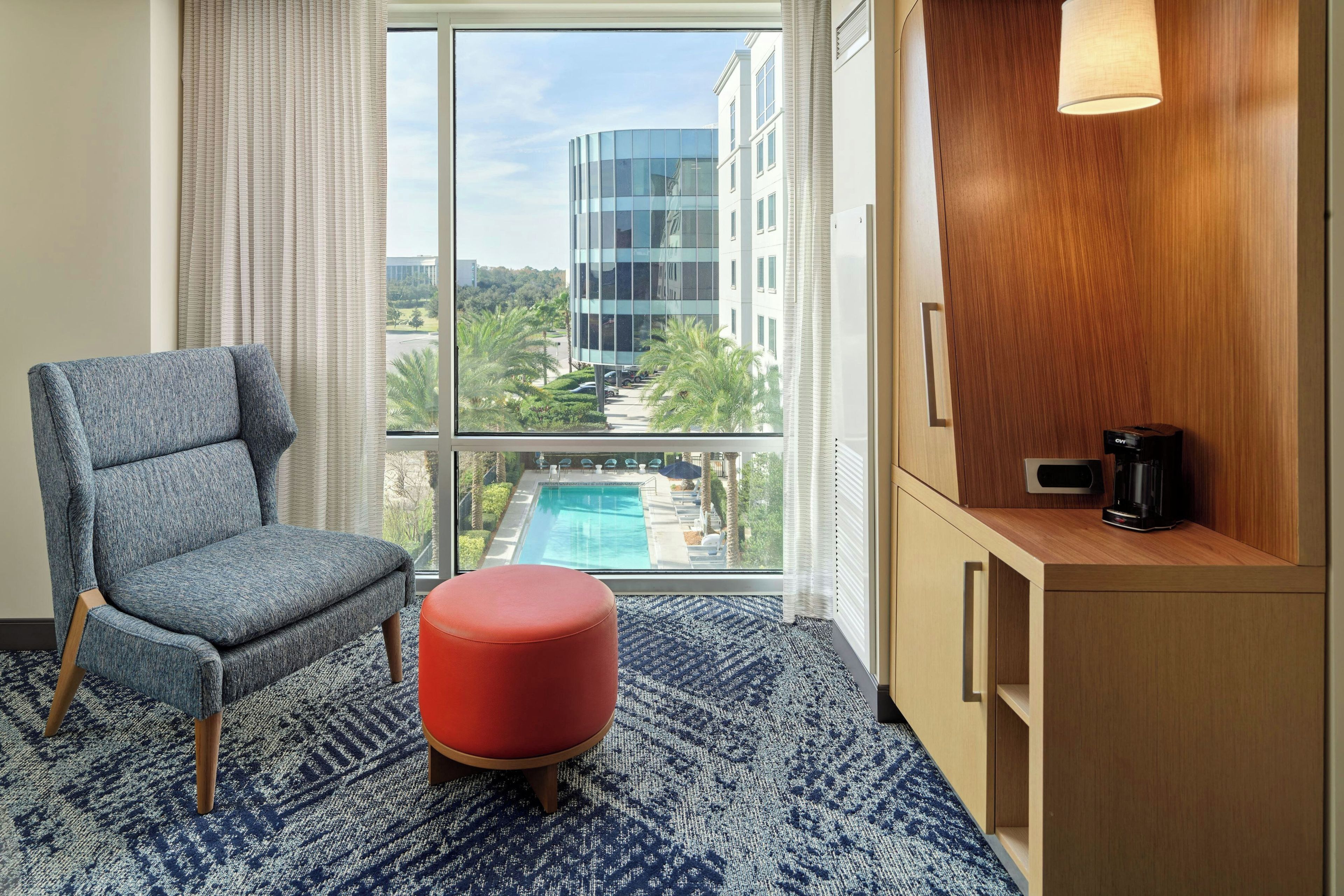 Une chambre d’hôtel dotée d’une grande fenêtre, d’un tapis à motifs bleus, d’un fauteuil gris, d’un pouf rouge, d’un buffet en bois avec une machine à café, et offrant une vue sur un bâtiment moderne et une piscine.