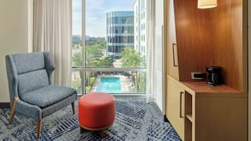 Une chambre d’hôtel dotée d’une grande fenêtre, d’un tapis à motifs bleus, d’un fauteuil gris, d’un pouf rouge, d’un buffet en bois avec une machine à café, et offrant une vue sur un bâtiment moderne et une piscine.