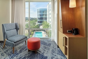 Ein Hotelzimmer mit einem großen Fenster, einem blau gemusterten Teppich, einem grauen Sessel, einem roten Hocker, einer Holzkommode mit einer Kaffeemaschine und Blick auf ein modernes Gebäude sowie einen Swimmingpool.
