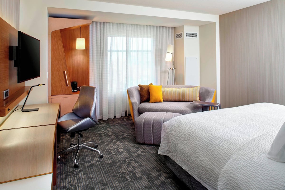Une chambre d’hôtel comprenant un lit, un bureau avec une chaise, un canapé avec des coussins, une télévision et une fenêtre avec des rideaux.
