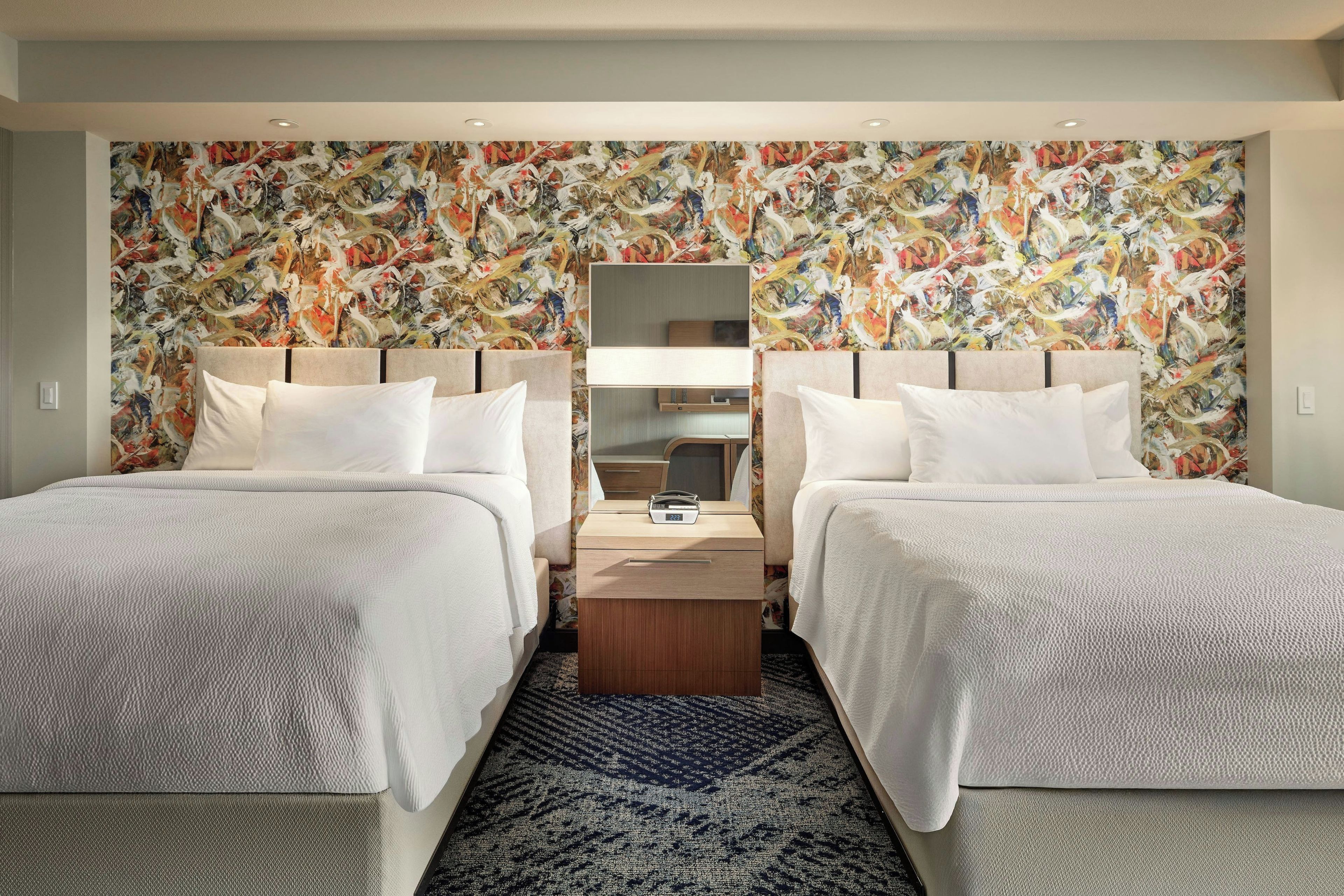 Een hotelkamer met twee bedden, een nachtkastje, een spiegel en een kleurrijk abstract schilderij.