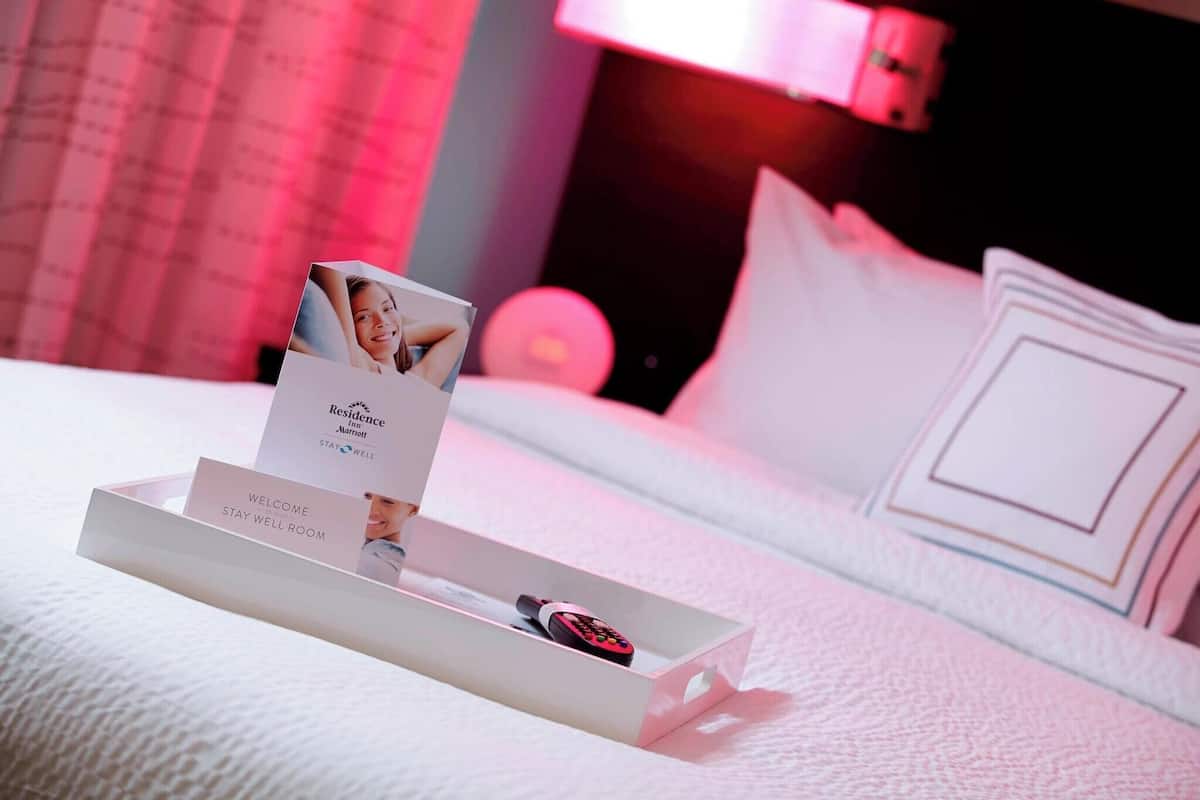 Un lit bien fait, recouvert d’une couette blanche, une table de chevet sur laquelle se trouve un plateau avec une carte de bienvenue, une télécommande et un coussin orné d’un motif géométrique.