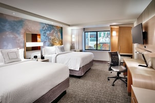A hotel room with two beds, a desk, a chair, a TV, and a large window with a view.