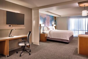 Habitación de hotel con televisión de pantalla plana, escritorio con silla, cama con ropa blanca y un ventanal con cortinas traslúcidas.