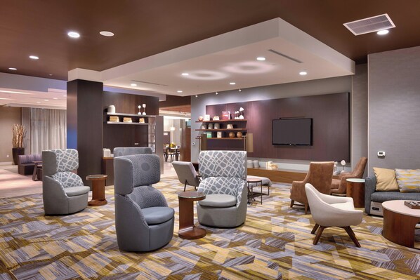 A modern hotel lobby with a variety of seating options, including armchairs and sofas, a patterned carpet, and a flat-screen TV mounted on the wall.