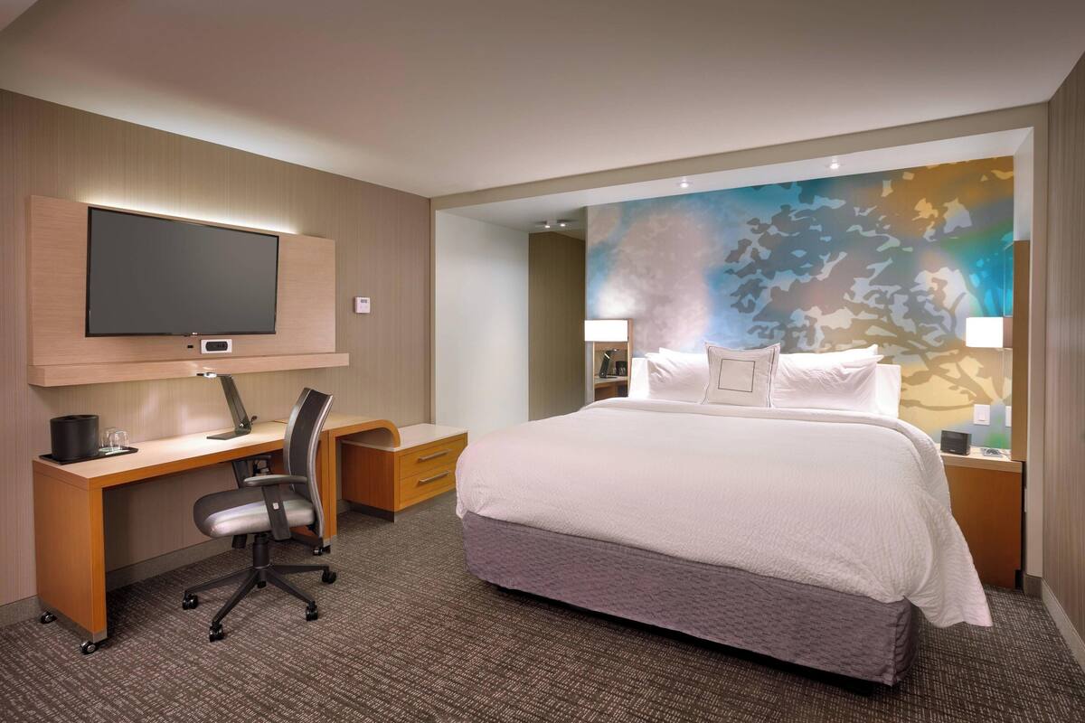 Ein Hotelzimmer mit Bett, Schreibtisch, Stuhl, Fernseher und einem großen abstrakten Gemälde an der Wand.