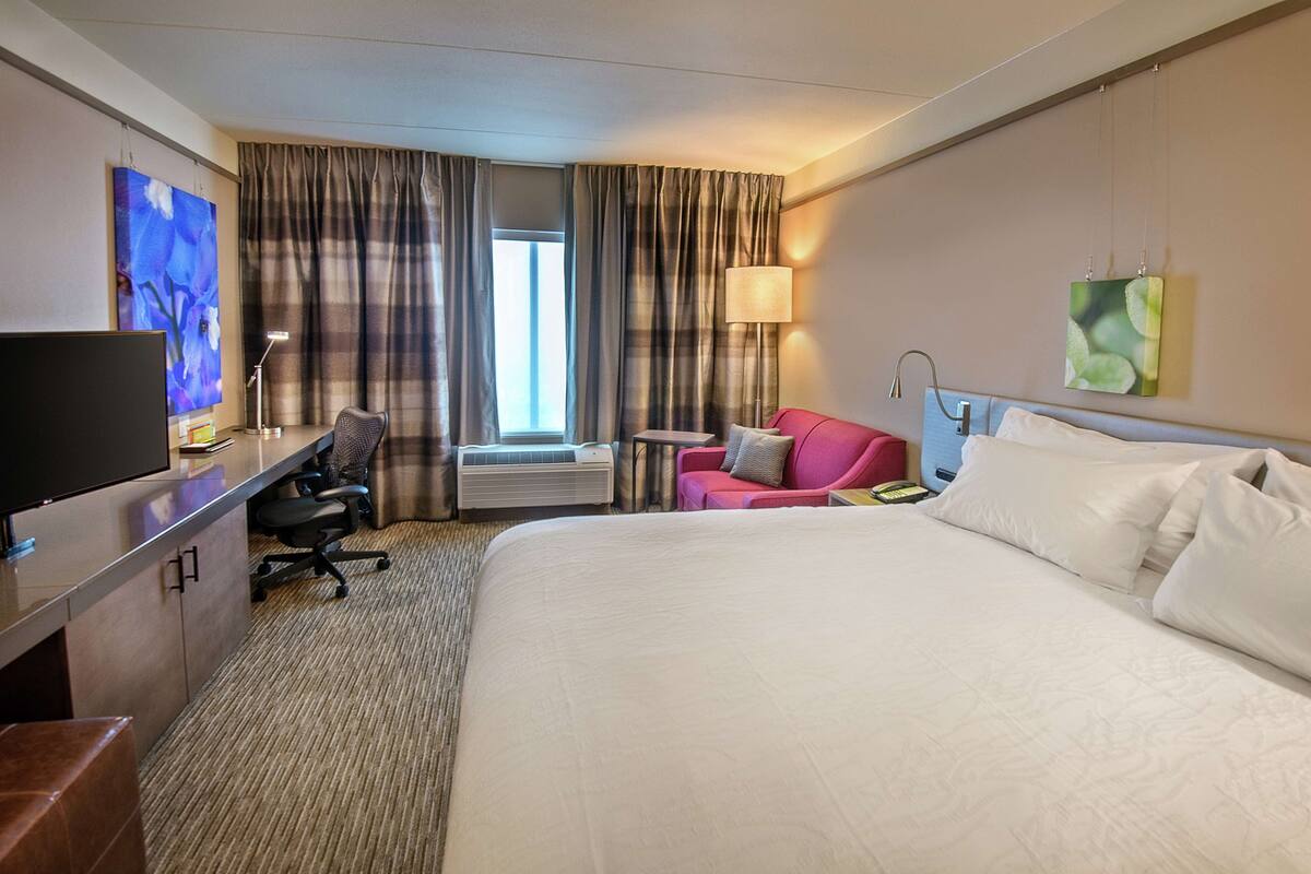 A hotel room with a bed, a desk with a computer, a chair, a TV, and a window with curtains.