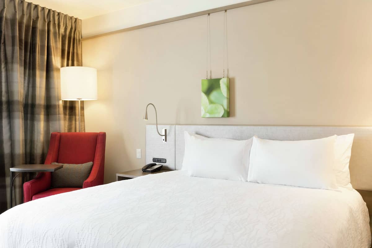 A hotel room with a large bed, a red armchair, a small table, a lamp, a telephone, and a green abstract painting on the wall.