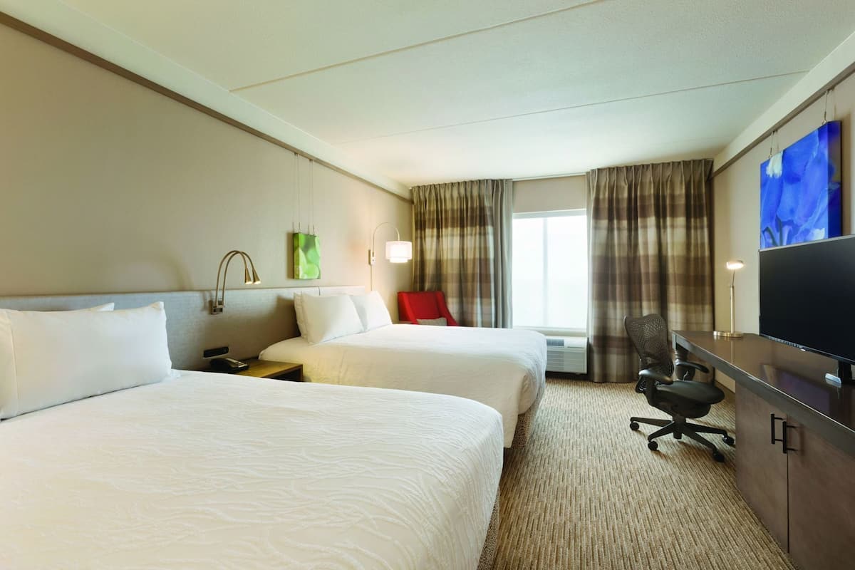 A hotel room with two beds, a desk with a TV, a chair, and a window with curtains.