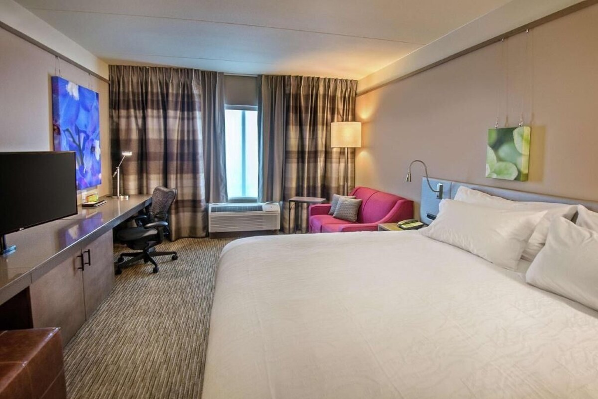 Habitación de hotel con cama, escritorio con televisor, silla, mesa pequeña y ventana con cortinas.
