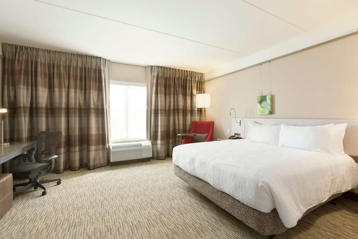 A hotel room with a large bed, a desk with a chair, a lamp, and a window with curtains.