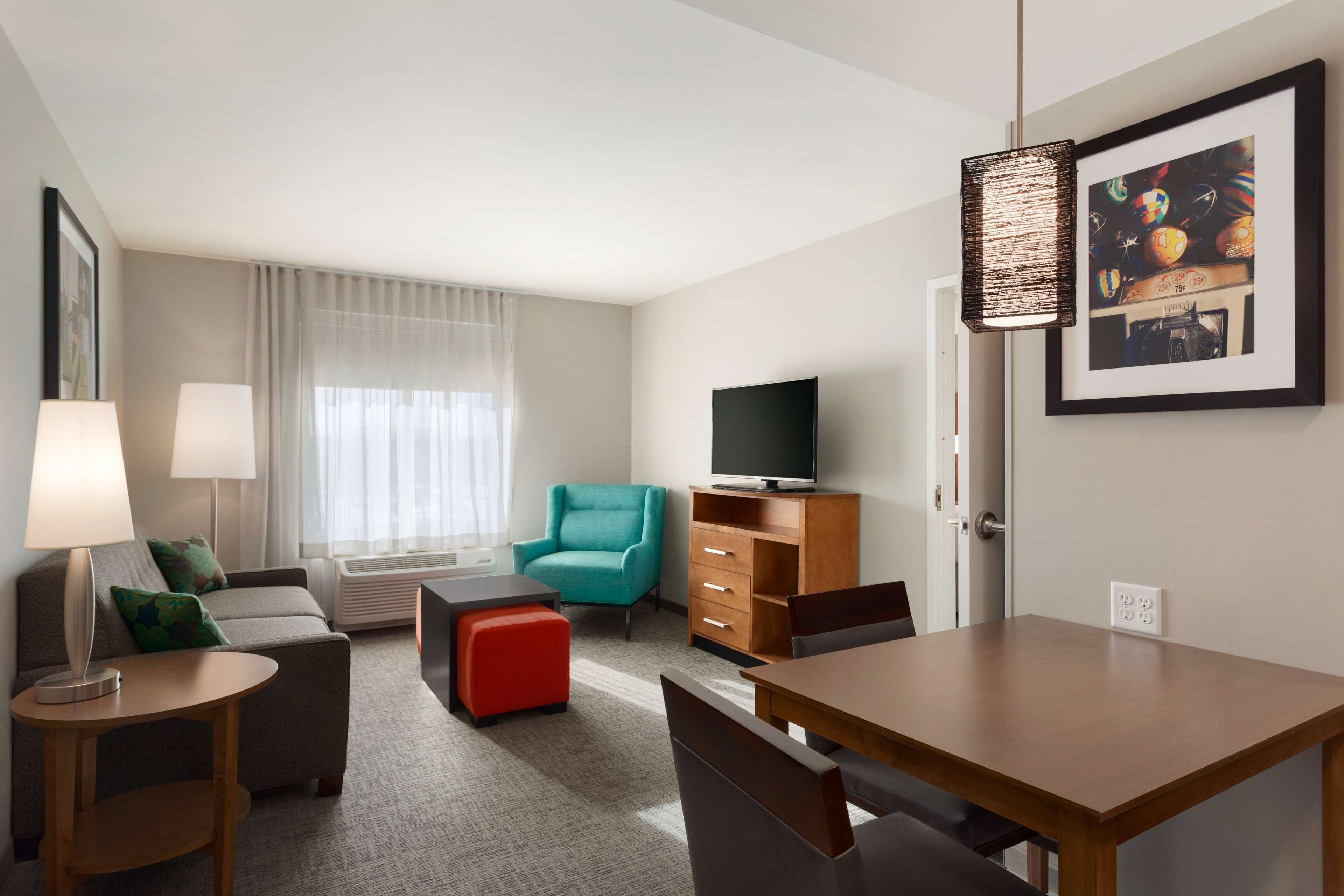 A hotel room with a sofa, a red ottoman, a teal armchair, a wooden table, a TV, and a framed picture on the wall.