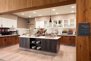 Een moderne keuken met houten kastjes, een centraal eiland met een spoelbak en de openingstijden van het ontbijt staan op de muur.