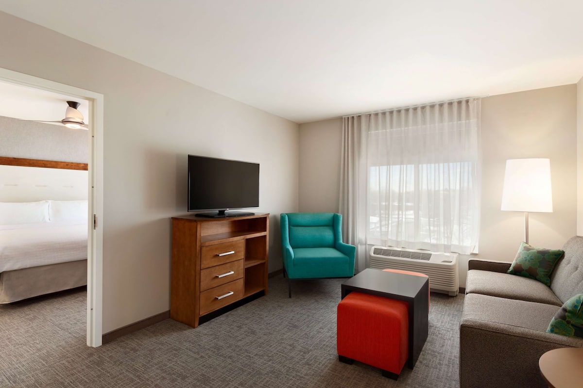 Habitación de hotel con una cama, un televisor sobre un aparador de madera, una silla verde azulada, un reposapiés rojo, un sofá gris con cojines verdes y una ventana con cortinas traslúcidas.