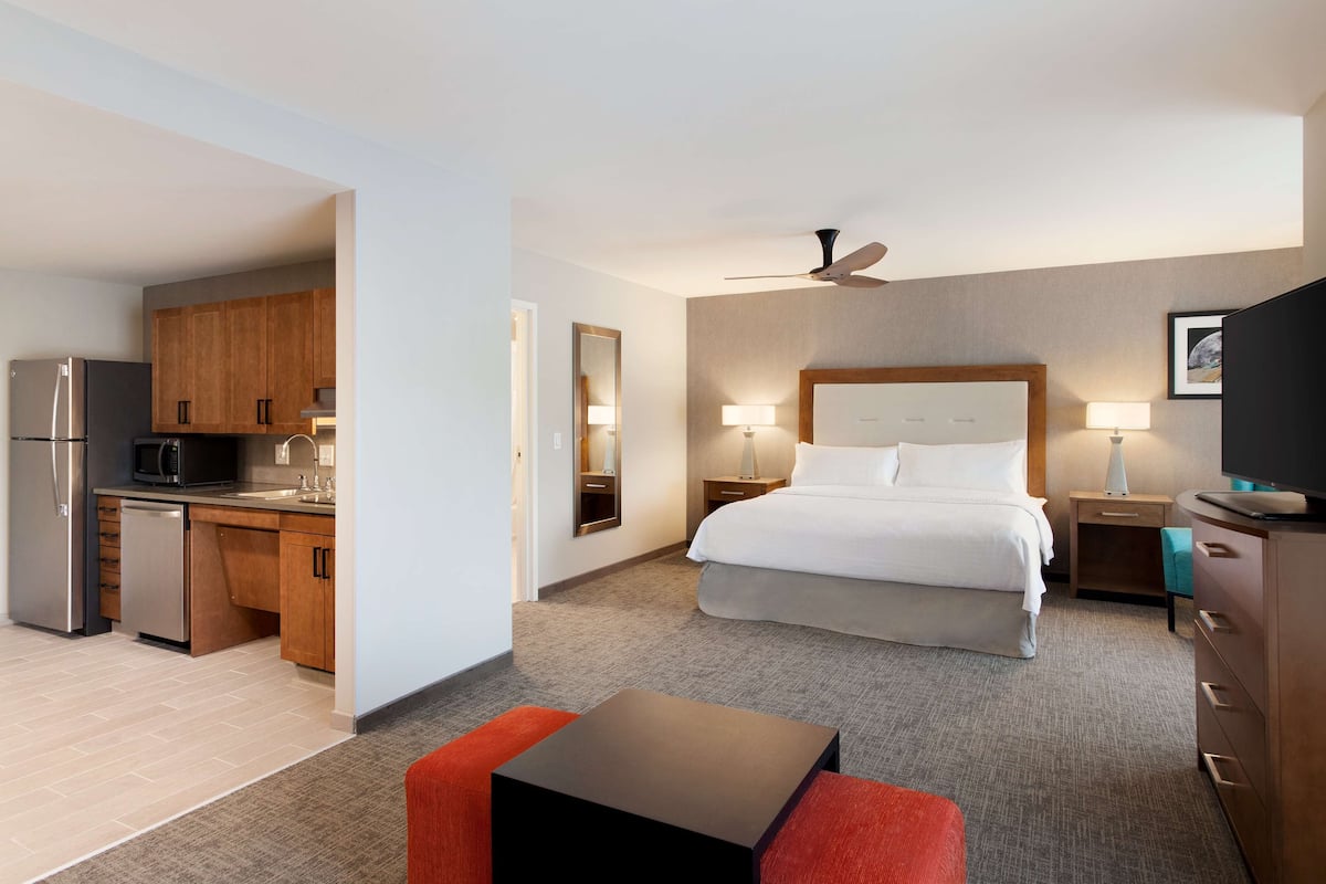 Habitación de hotel con cama, mesitas de noche, escritorio con silla, kitchenette y ventilador de techo.
