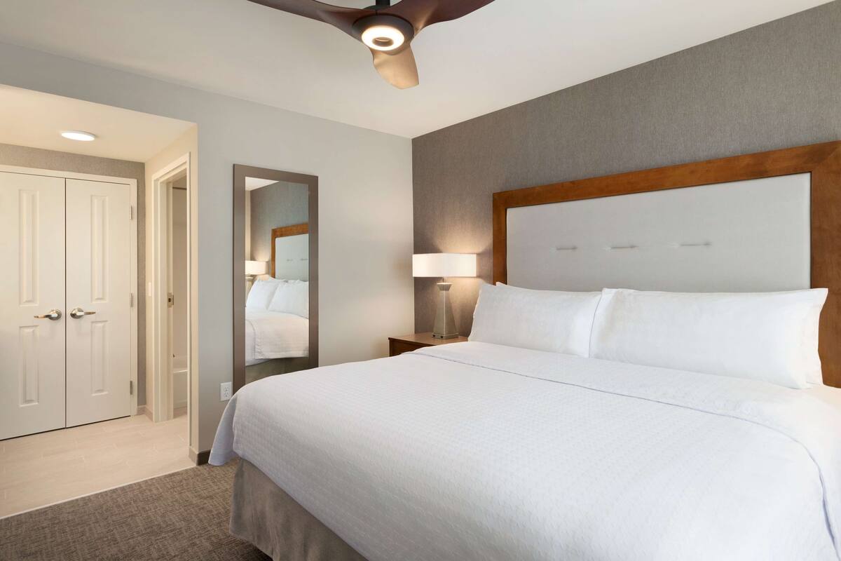 Habitación de hotel con una cama grande, un ventilador de techo, una mesita de noche con lámpara y una puerta que da a otra habitación.