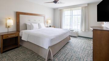 A hotel room with a large bed, two bedside tables with lamps, a ceiling fan, a large window with curtains, and a TV mounted on the wall.