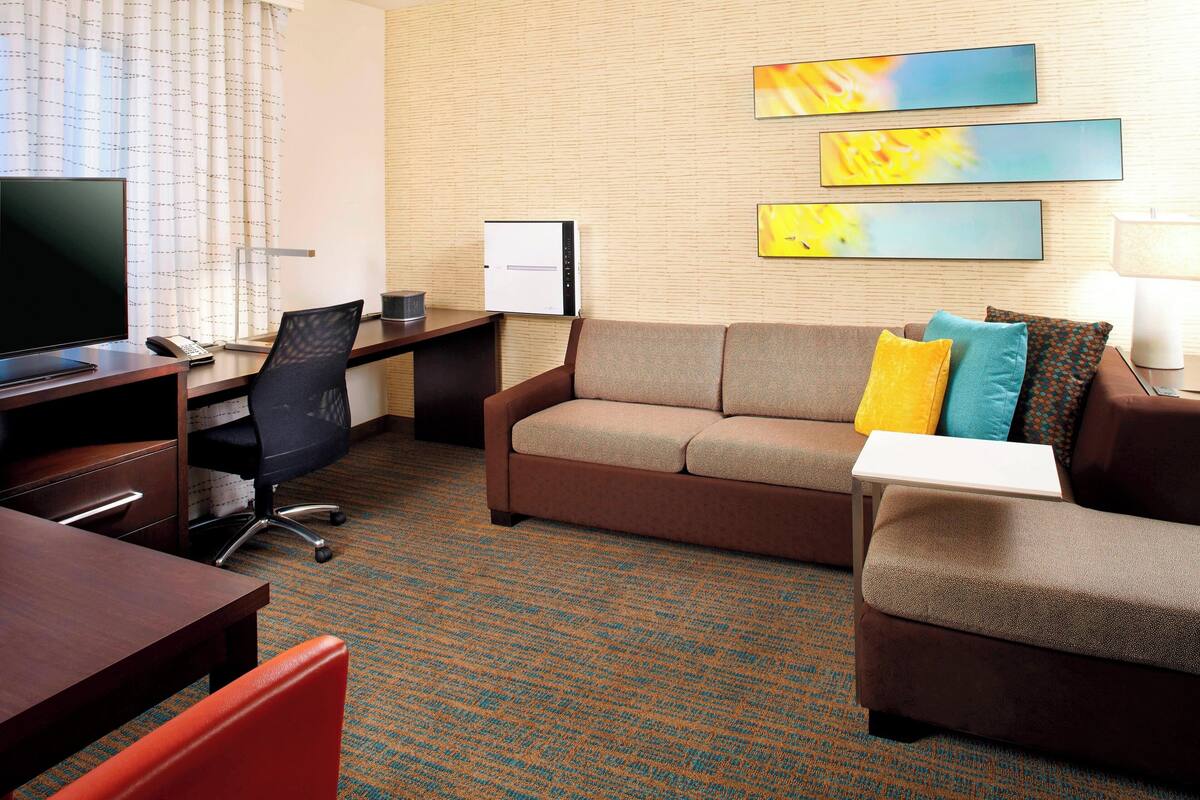 Habitación de hotel con un sofá marrón, un escritorio con una computadora, un televisor y un monitor de pantalla plana. Hay tres cuadros abstractos en la pared.