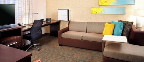 Een hotelkamer met een bruine bank, een bureau met een computer, een televisie en een flatscreenmonitor. Er hangen drie abstracte schilderijen aan de muur.