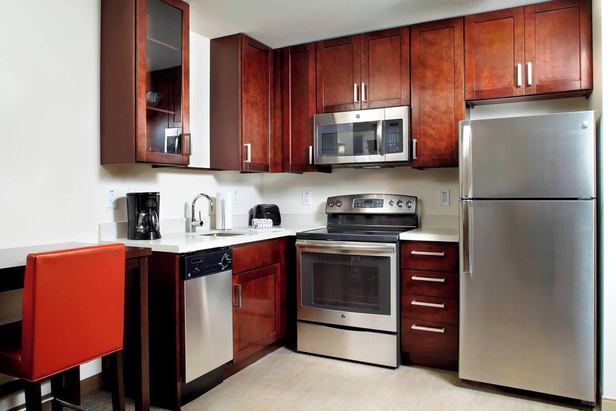 Cocina compacta con armarios de madera, electrodomésticos de acero inoxidable, incluyendo microondas, horno y refrigerador, además de una pequeña zona de comedor con una silla roja.