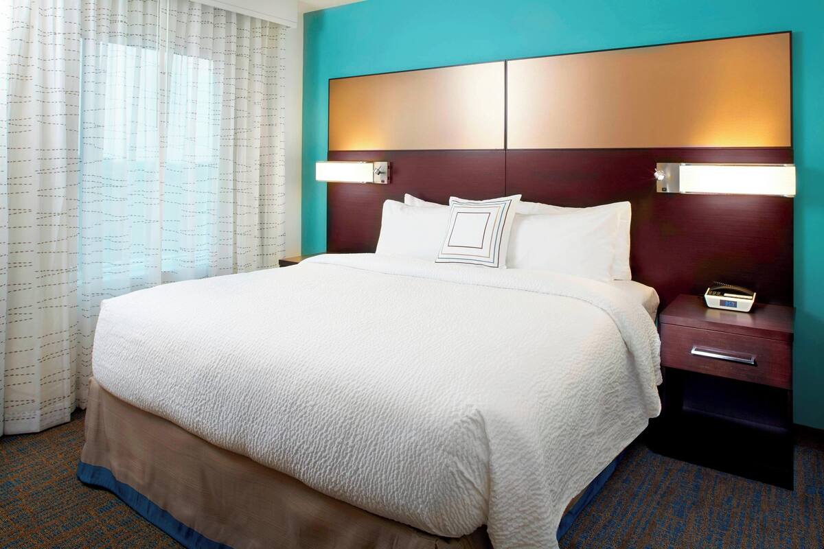 Habitación de hotel con una cama grande, una mesita de noche con lámpara y vista a la ciudad a través de la ventana.