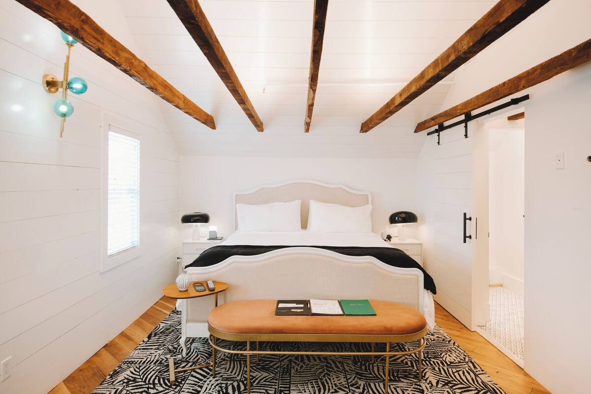 Une chambre à coucher avec un lit, des tables de chevet, un banc, une fenêtre et une porte.