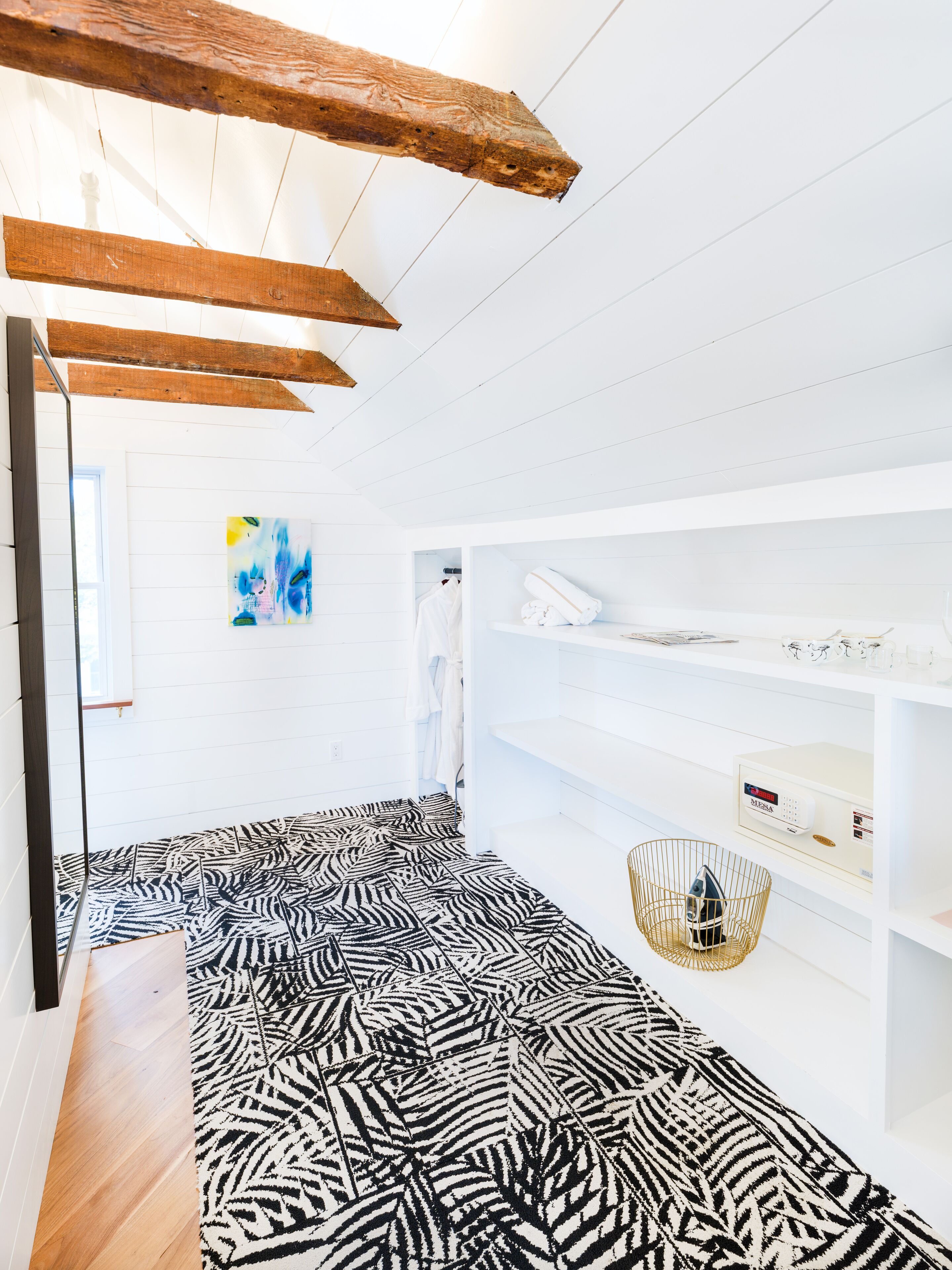 Une pièce avec un tapis à motifs zébrés, des poutres en bois au plafond, un mur blanc orné d’un tableau abstrait coloré et une étagère blanche sur laquelle sont disposés divers objets.