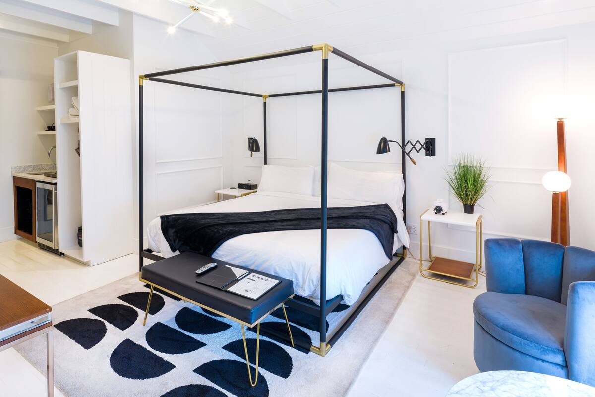 Une chambre d’hôtel moderne avec un lit à baldaquin, un petit bureau, un fauteuil bleu et une plante en pot.