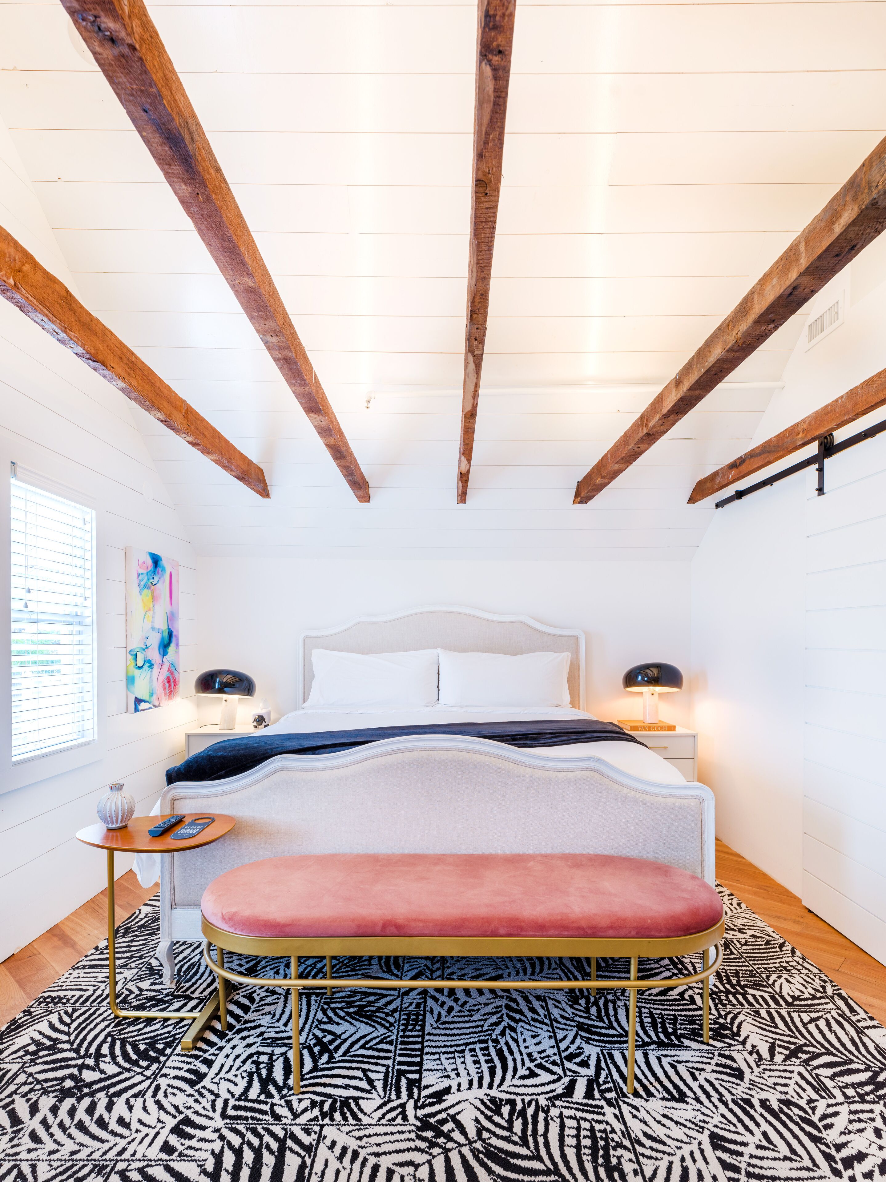 A bedroom with a bed, bedside tables, a bench, a zebra-patterned rug, and exposed wooden beams.