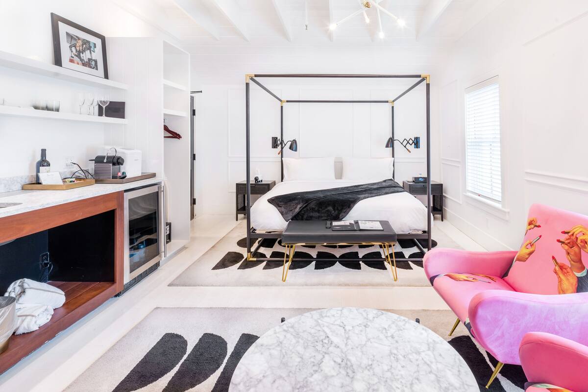 Une chambre d’hôtel moderne avec un lit à baldaquin, un bureau avec un ordinateur, un fauteuil rose et une table basse en marbre.