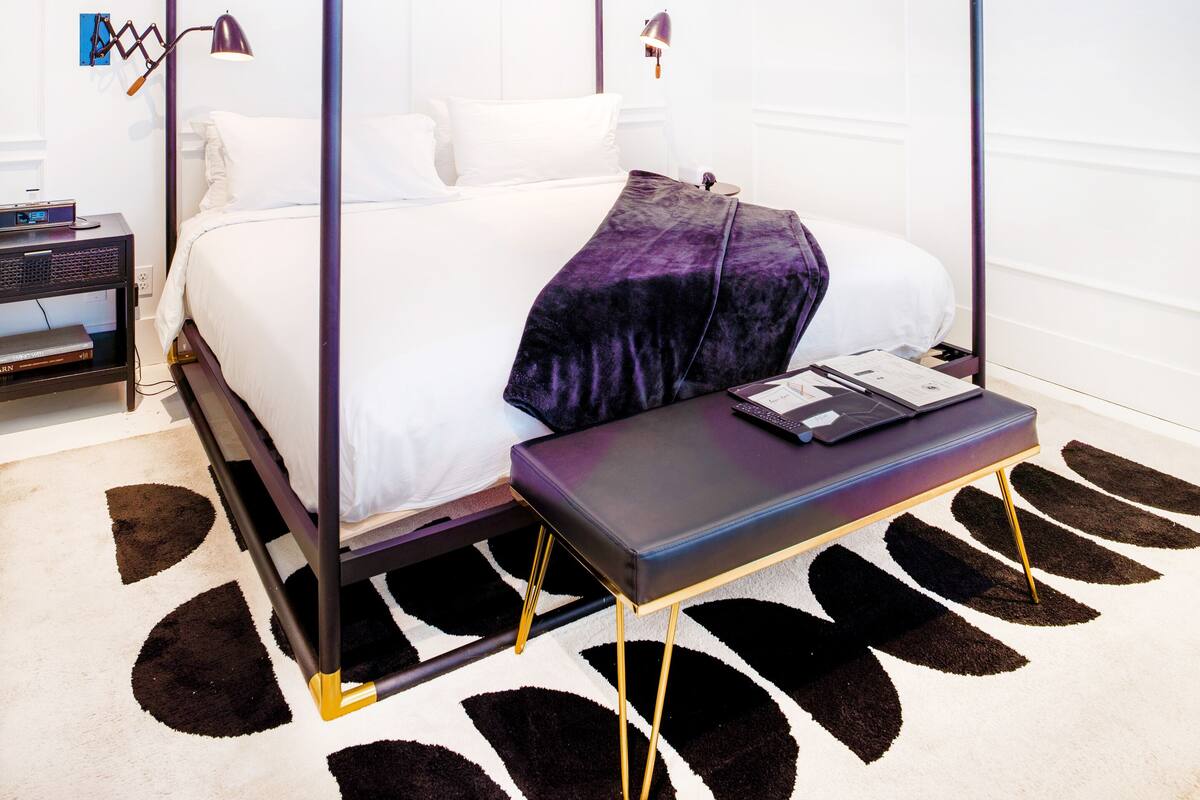 Un lit à baldaquin à quatre poteaux, agrémenté d’un tapis à motifs noir et blanc, d’une petite table de chevet et d’une table de nuit avec une lampe.