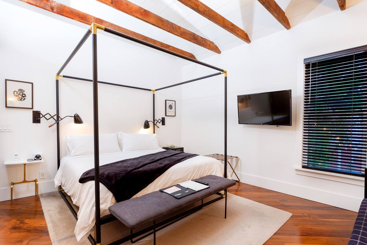 Un lit à baldaquin avec une banquette, des tables de chevet, un téléviseur à écran plat et une fenêtre avec des stores.