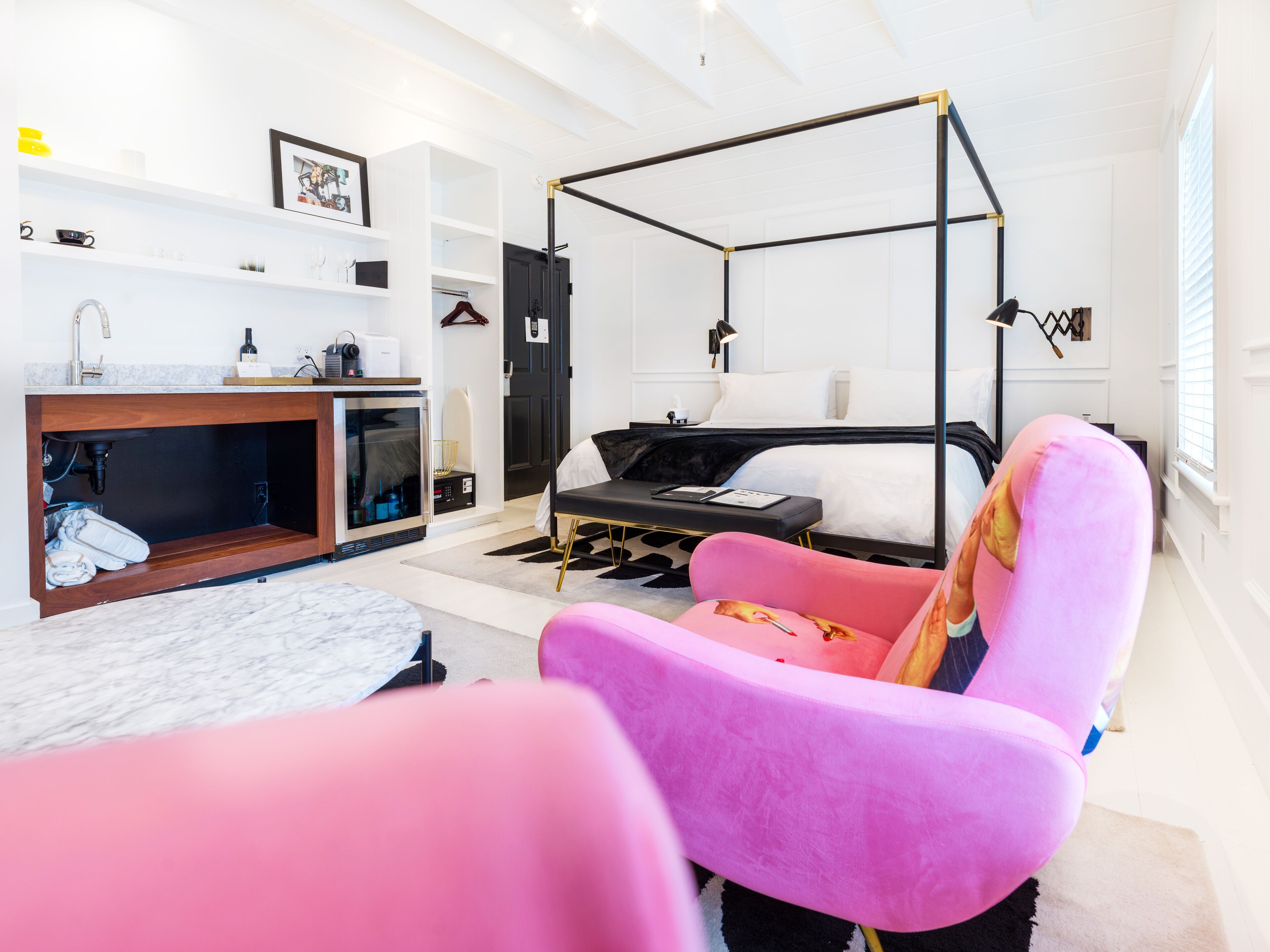 Un dormitorio moderno con cama con dosel, tocador con lavabo, vestidor y una silla rosa.