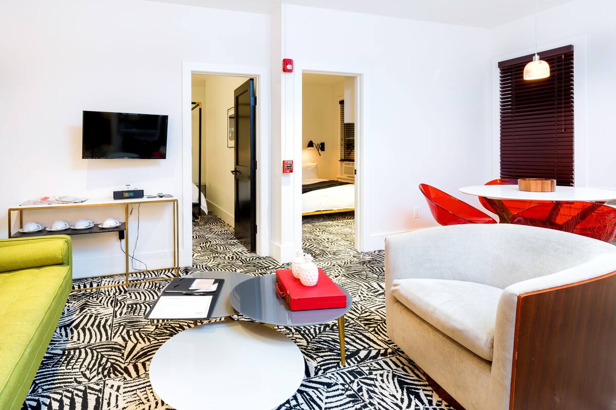 Une chambre d’hôtel moderne avec un sol à motifs zébrés, un téléviseur à écran plat, un canapé, un fauteuil rouge et une petite table sur laquelle se trouve un bol.