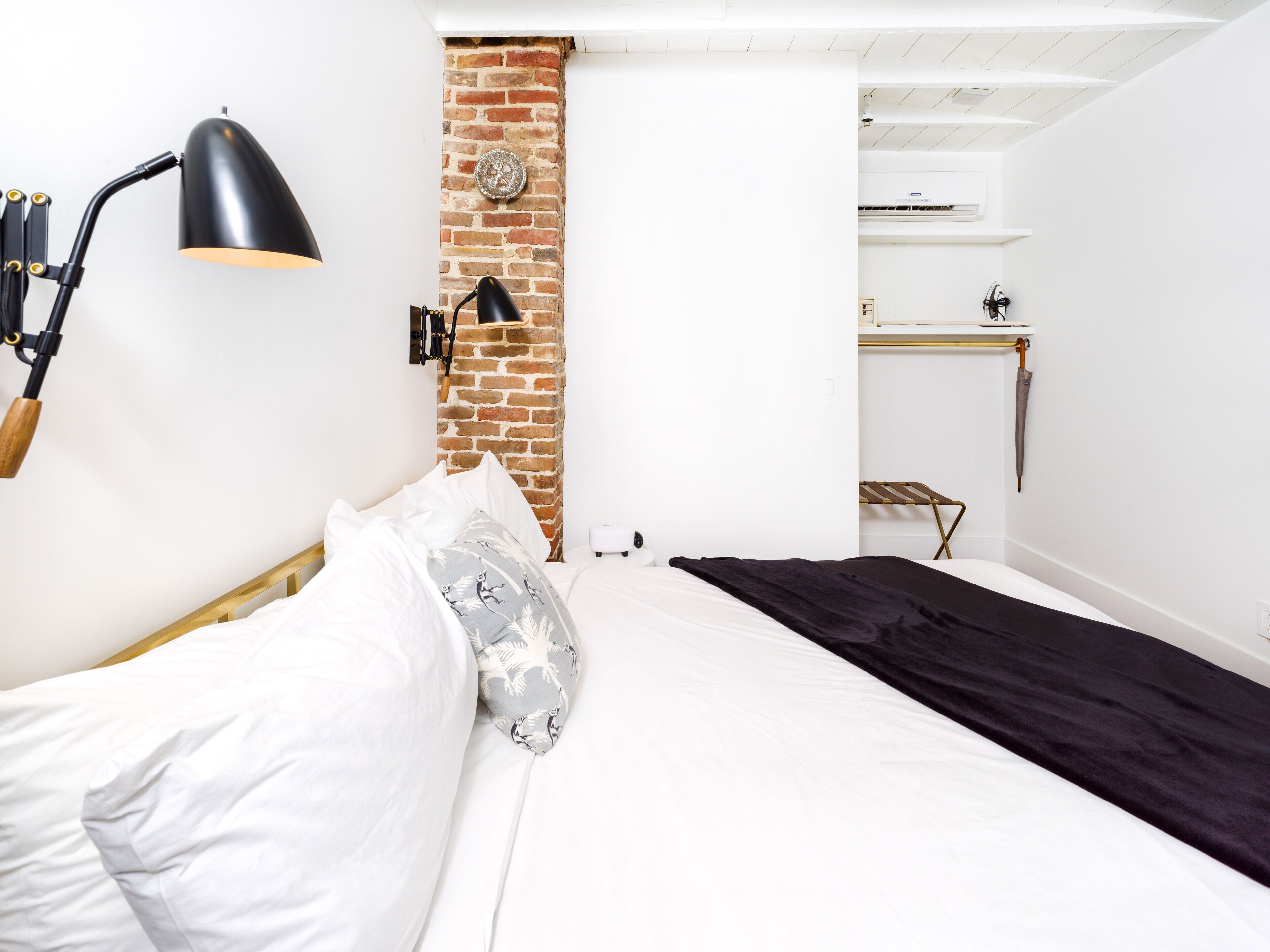 Un dormitorio con pared de ladrillo, cama con ropa de cama blanca y negra, lámpara de escritorio negra y una pequeña mesa con una silla.